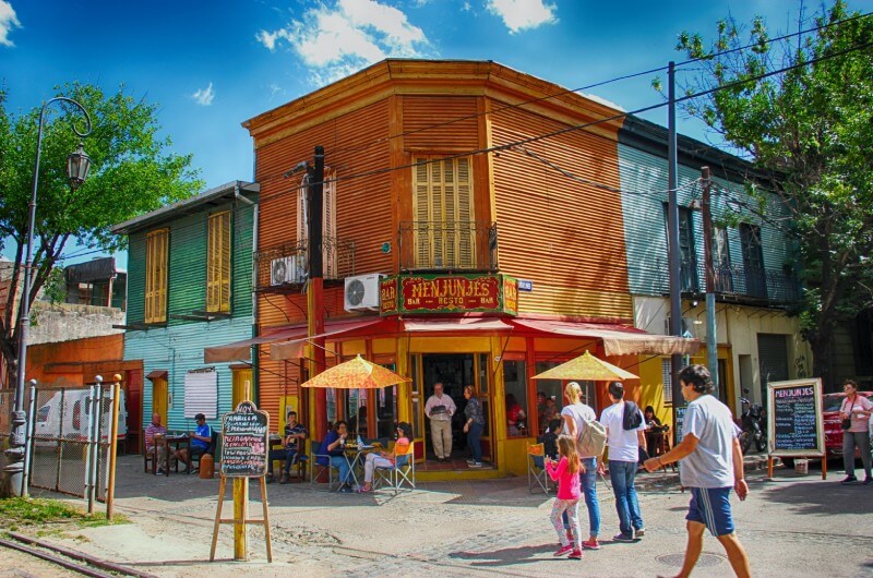 A photo of a colorful street corner with a restaurant and people walking by. The restaurant is a two-story wooden building with a yellow and orange exterior and a sign that reads “MEN JUNJES”. There are people sitting at tables with umbrellas on the sidewalk in front of the restaurant and people walking on the sidewalk and crossing the street. The background consists of a blue sky with clouds and trees.